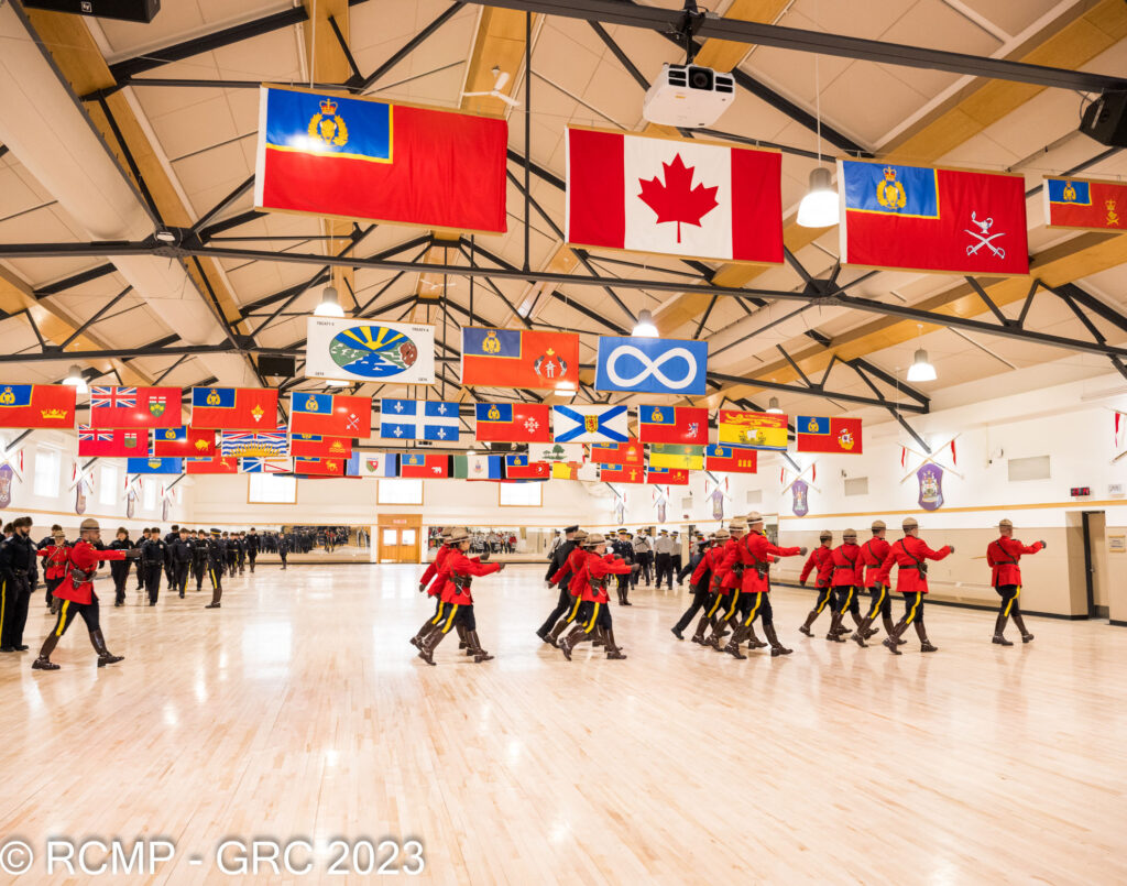 RCMP Cadets in Red Serge, marching in the hall at Depot with flags above.