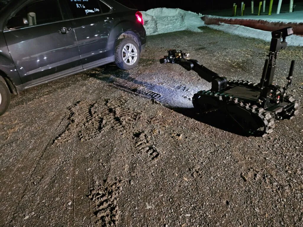 A modern-day robot approaching a vehicle containing potential explosives. It is remotely operated by an EDU Member who is positioned safe distance.